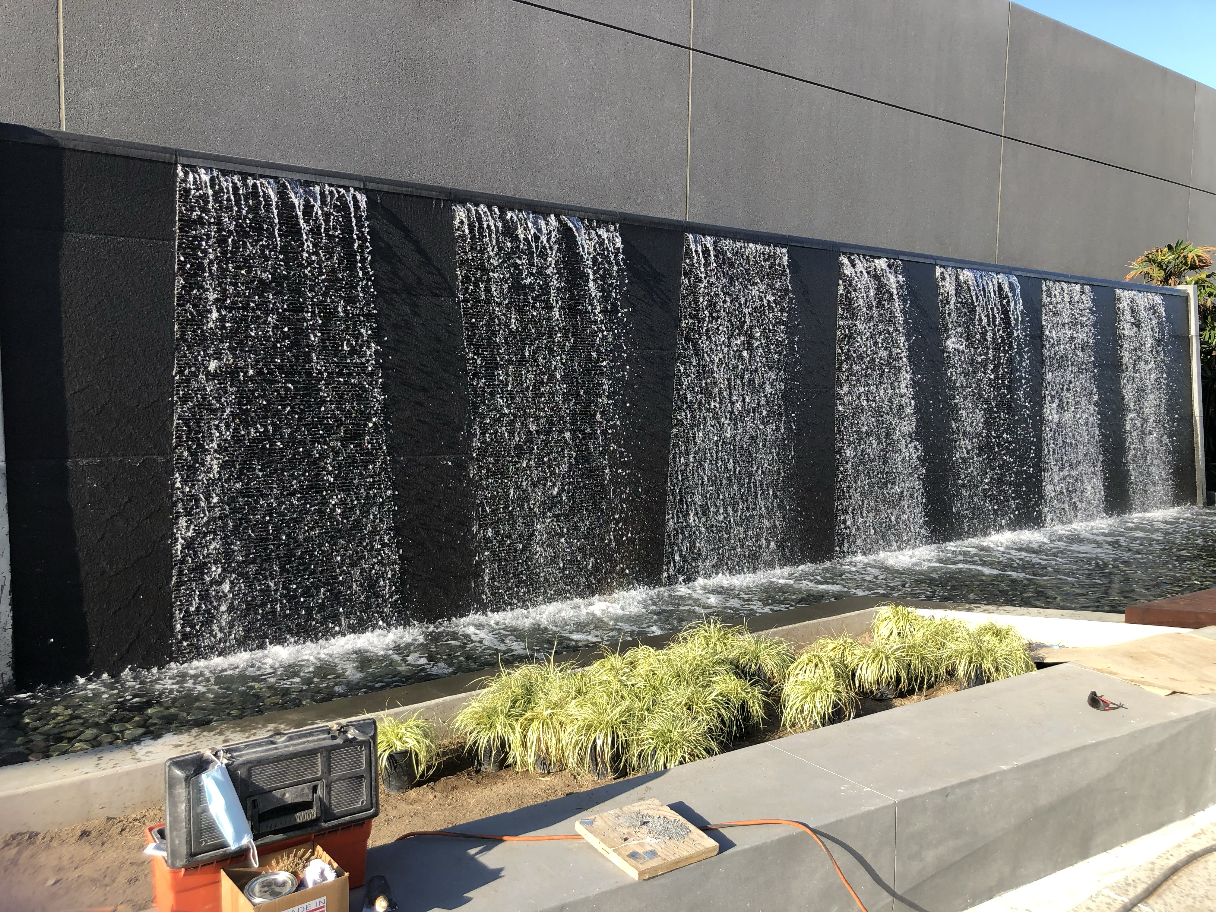 Modern water feature with multiple waterfalls cascading down a dark wall into a pebble pool.